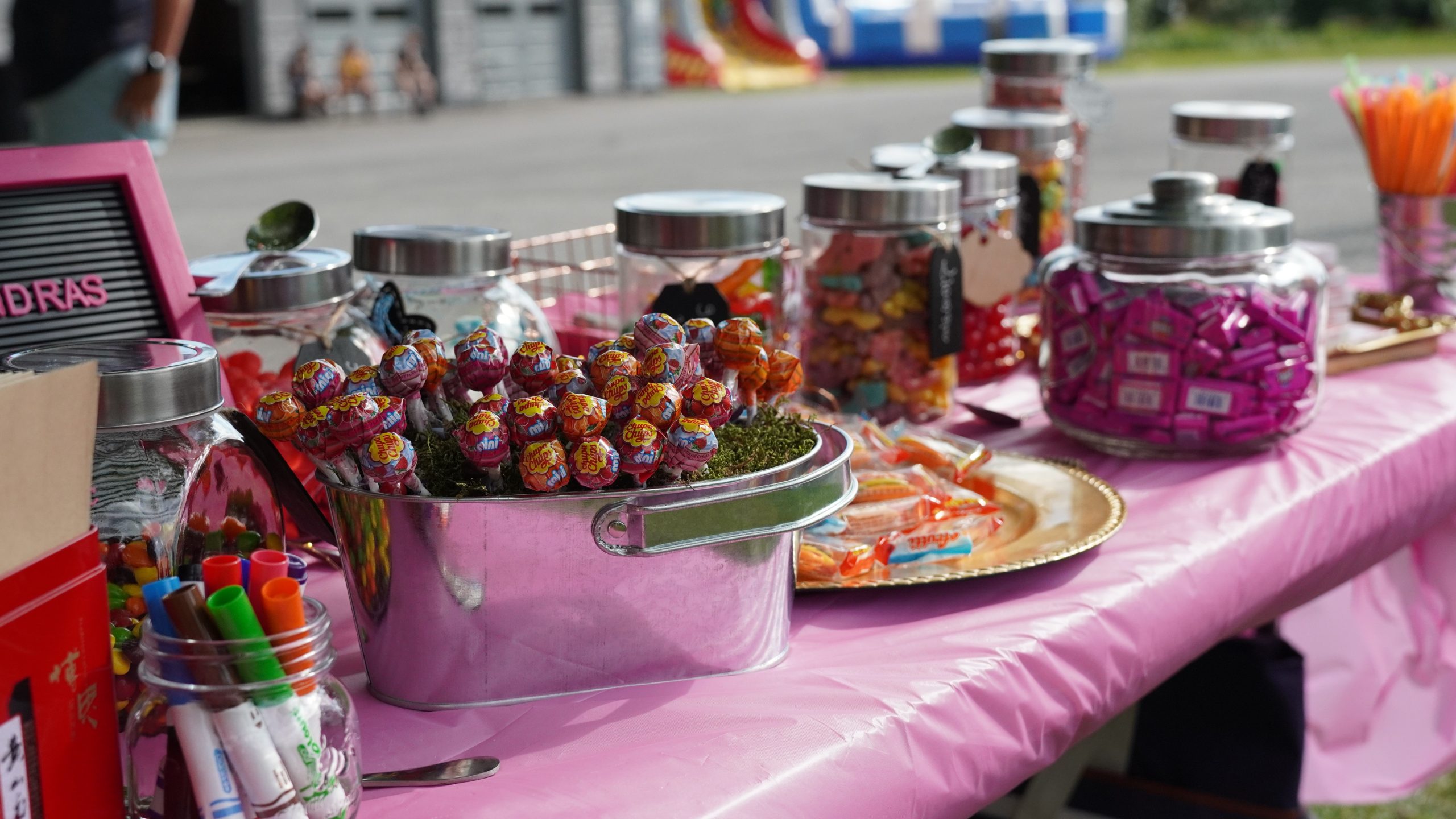 A table covered with a pink tablecloth holds jars of assorted candy, a bucket of lollipops, snacks on a tray, and various colorful markers.