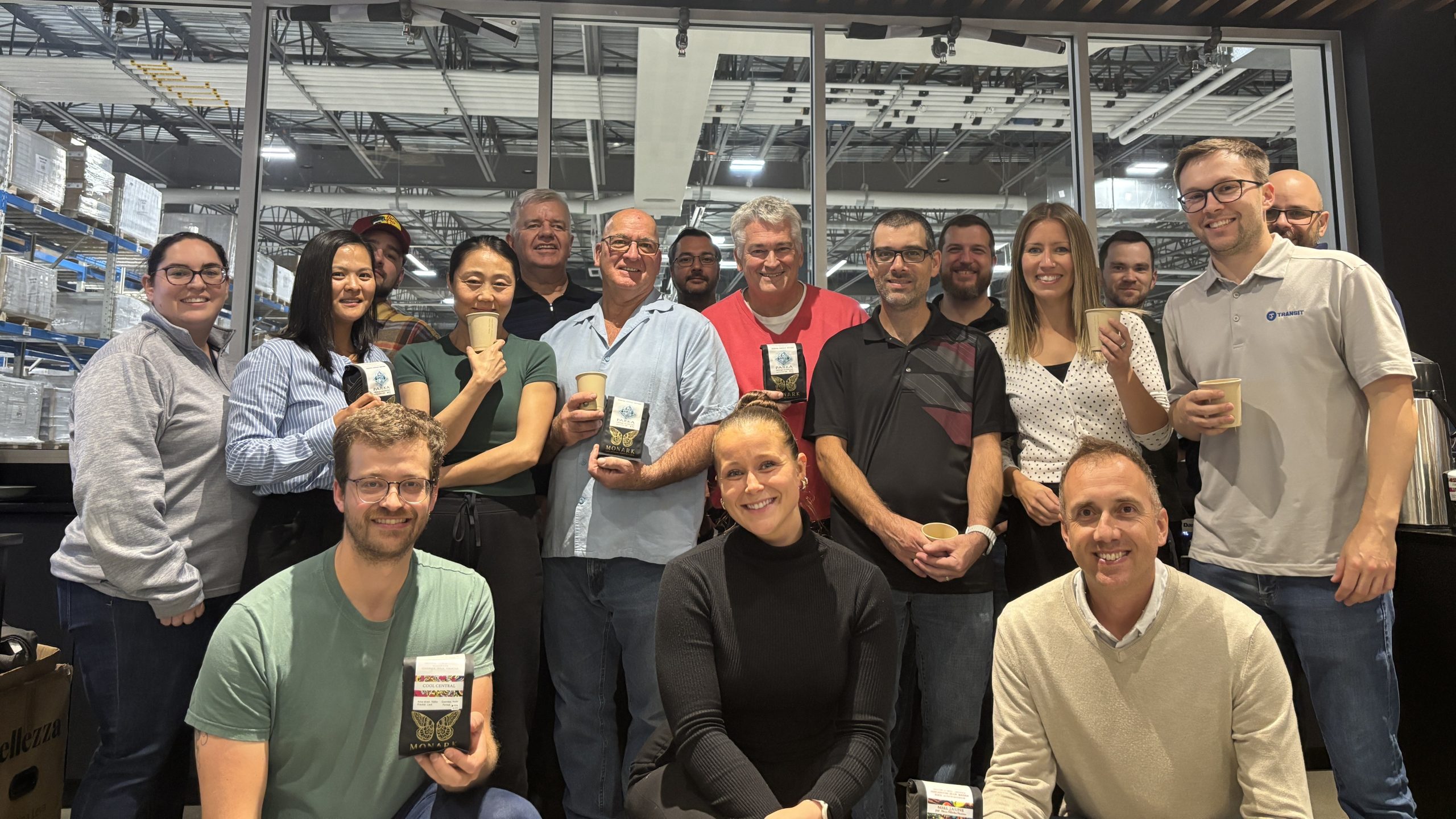 A group of adults poses indoors, some holding coffee cups and packaged items, with an industrial backdrop featuring large windows and visible ceiling structures.