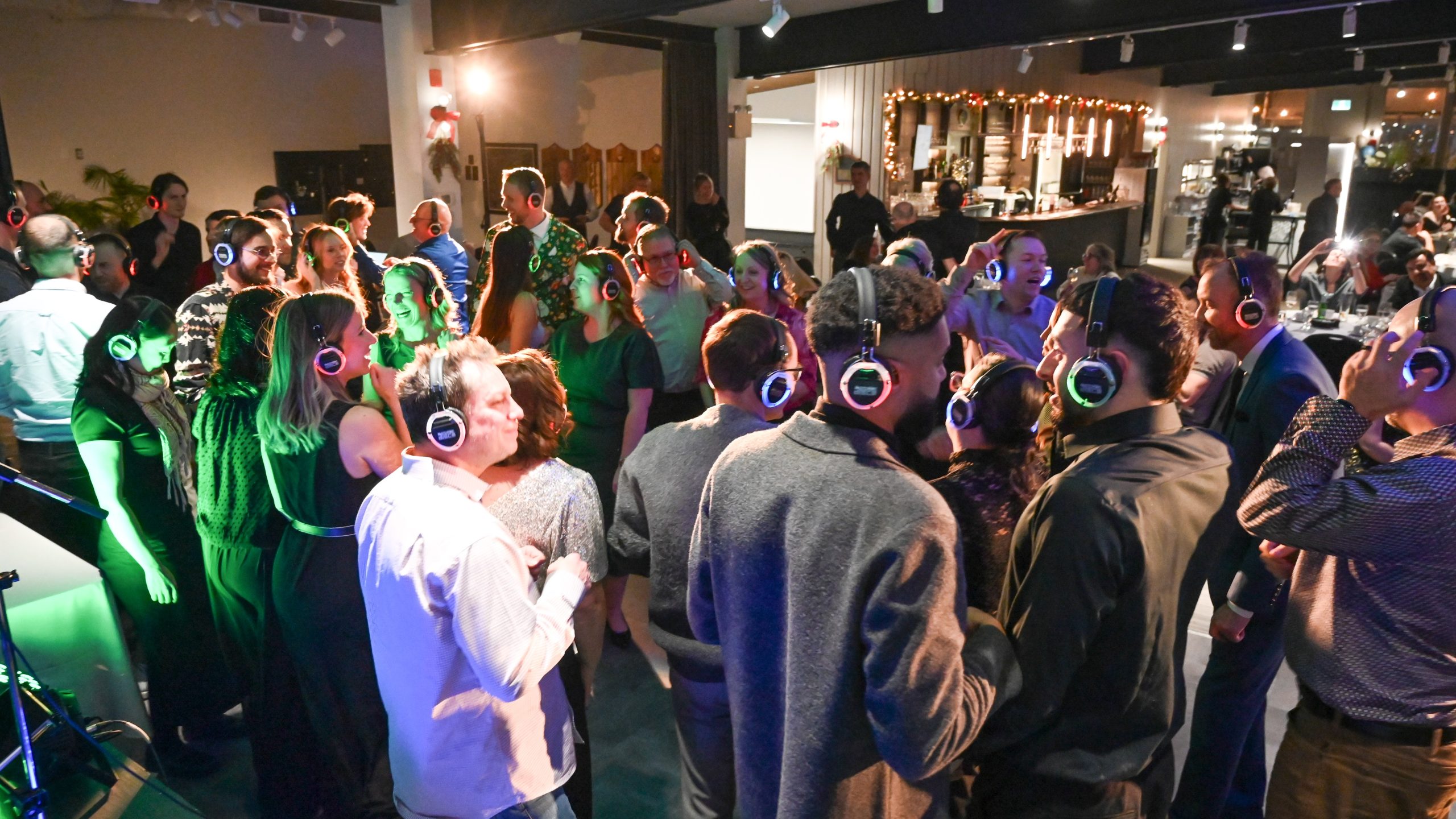 A group of people wearing wireless headphones dance and socialize at an indoor silent disco party, with a bar visible in the background.