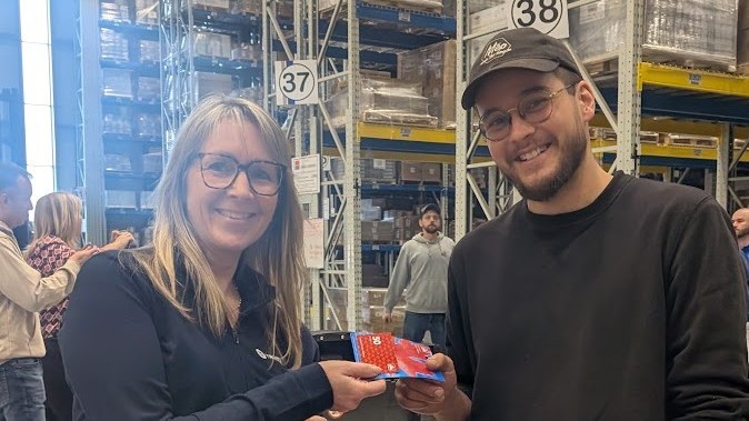 Two people stand in a warehouse; one hands a red card to the other. Both are smiling and wearing glasses. Shelves of boxes and numbered signs are visible in the background.
