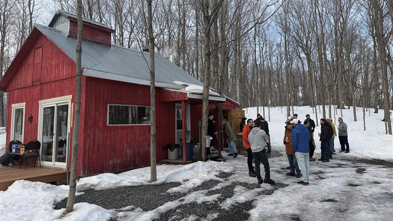 A group of people stands outside a small red wooden building surrounded by leafless trees and snow on the ground.