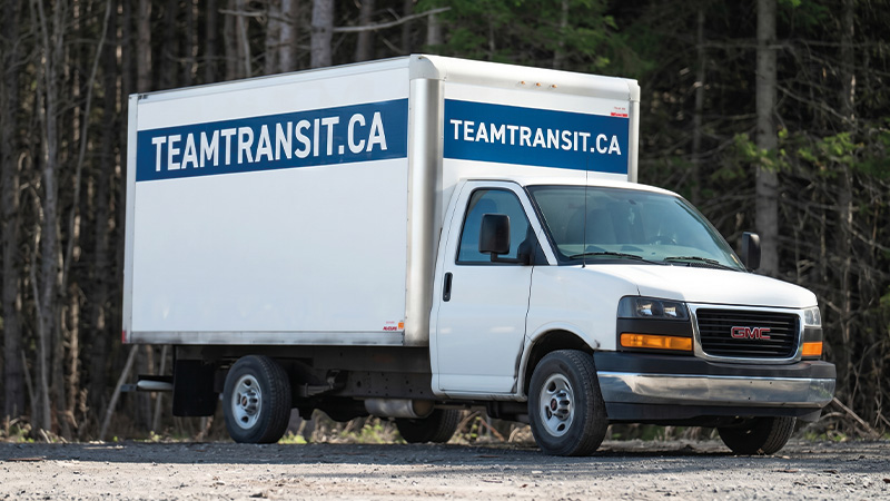 A white GMC box truck with TEAMTRANSIT.CA in blue text is parked on a gravel surface near a forested area.