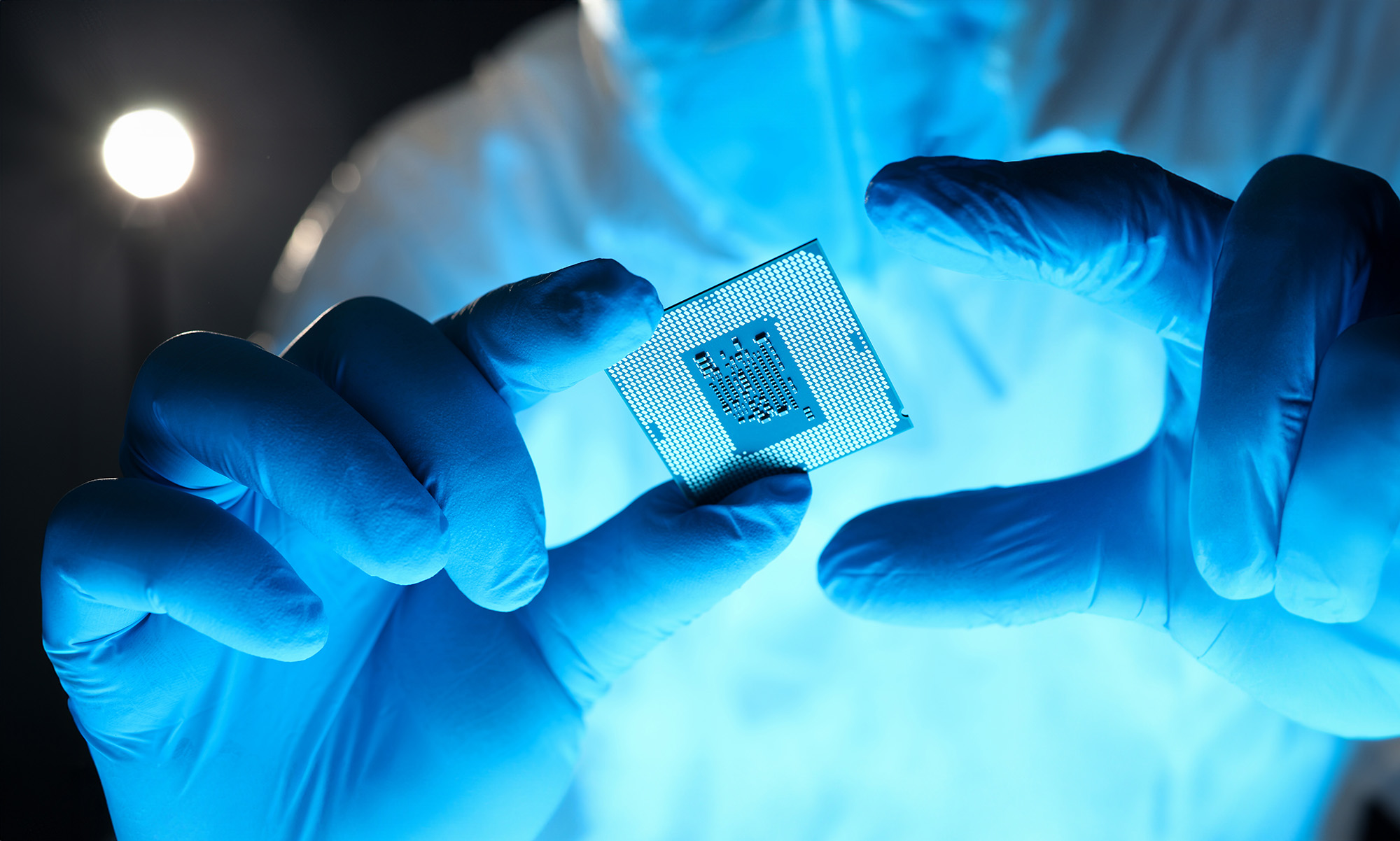 A person in blue gloves and a protective suit holds a computer microchip between their fingers, with a bright light source in the background.