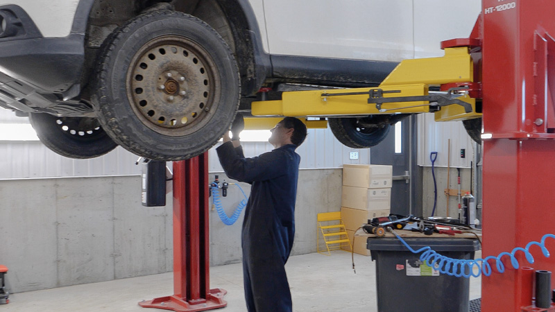 A mechanic in coveralls works on a white car elevated on a hydraulic lift in an auto repair shop.