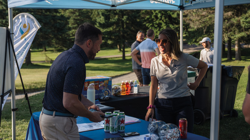Two people talk at an outdoor event booth under a canopy. Drinks, snacks, and promotional items are displayed on the table. Other people stand in the background near a grassy area.